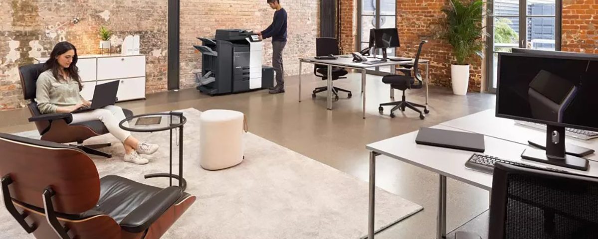 Ufficio moderno con pareti in mattoni, donna al computer portatile e uomo che utilizza una stampante multifunzione.