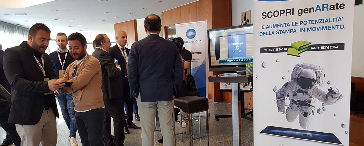 Persone in abbigliamento formale e casual a una fiera tecnologica con stand GenARate e Konica Minolta, mostra di innovazioni digitali e AR.